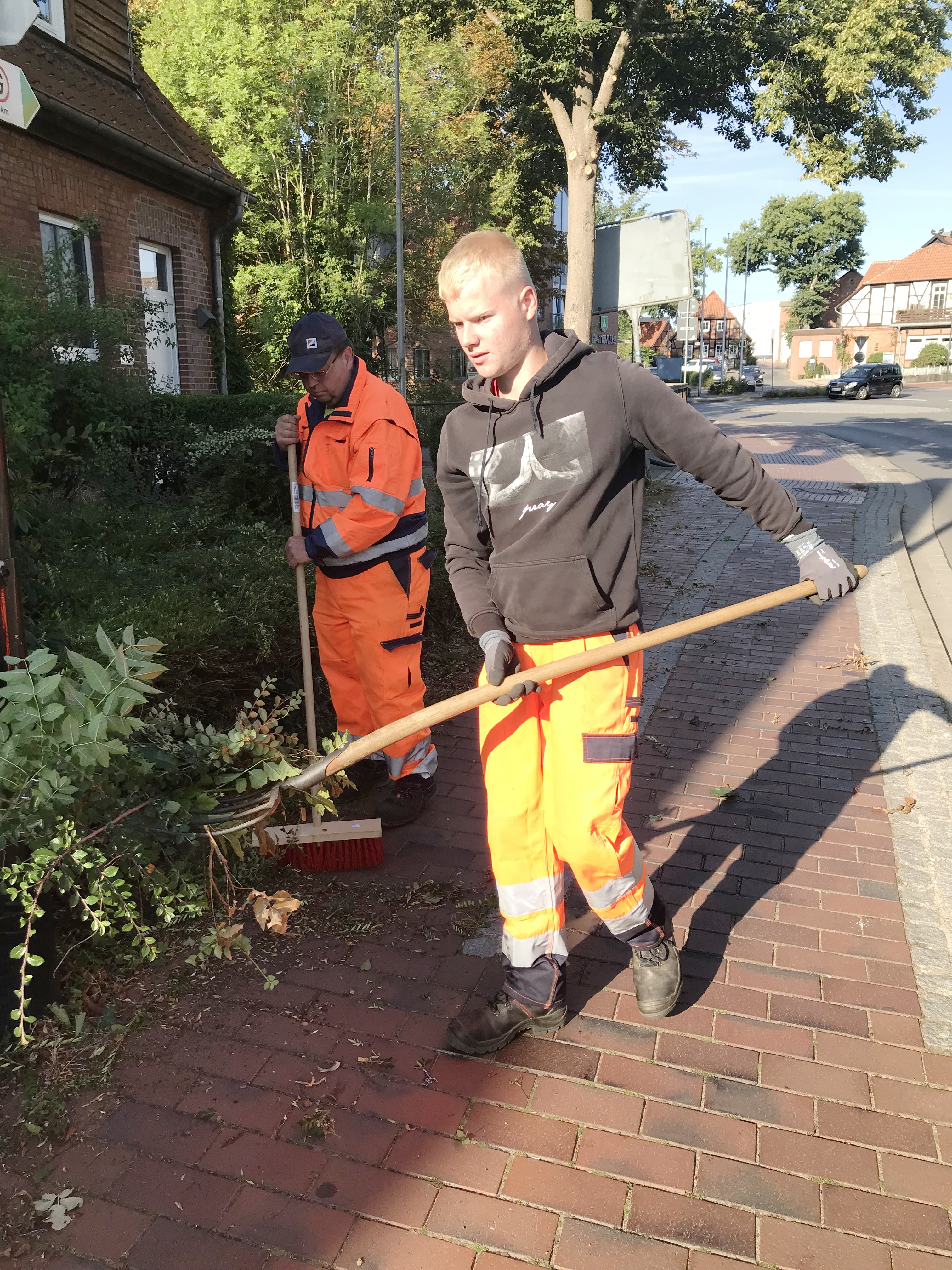 Vischer und Schröder: Arne Schröder (r.) trägt den Grünschnitt, den Erny Vischer bereits zusammen-gefegt hat, zu einer Schubkarre. Foto: Gemeinde Bienenbüttel